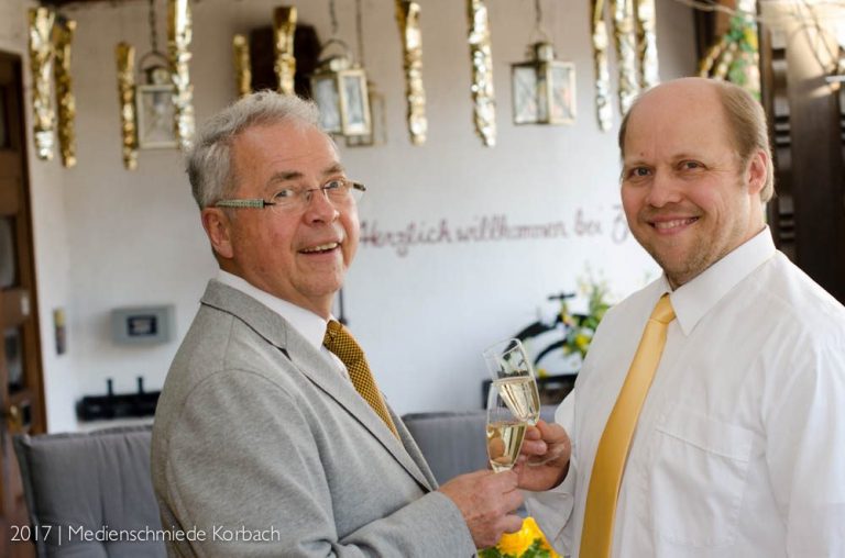Inhaber Uli und Urs Zabel Zwei Männer in Anzügen lächeln und stoßen mit Gläsern an, festliche Dekoration im Hintergrund.
Inhaber Uli und Urs Zabel